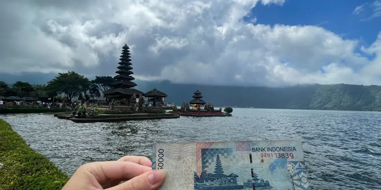 Ręka trzyma banknot 50 000 rupii na tle świątyni na wodzie. To symboliczne, że wiza turystyczna na Bali jest w zasięgu ręki!