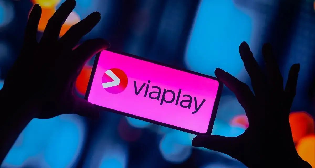Dłonie trzymają smartfon z logo Viaplay. W tle rozmyte, kolorowe światła.