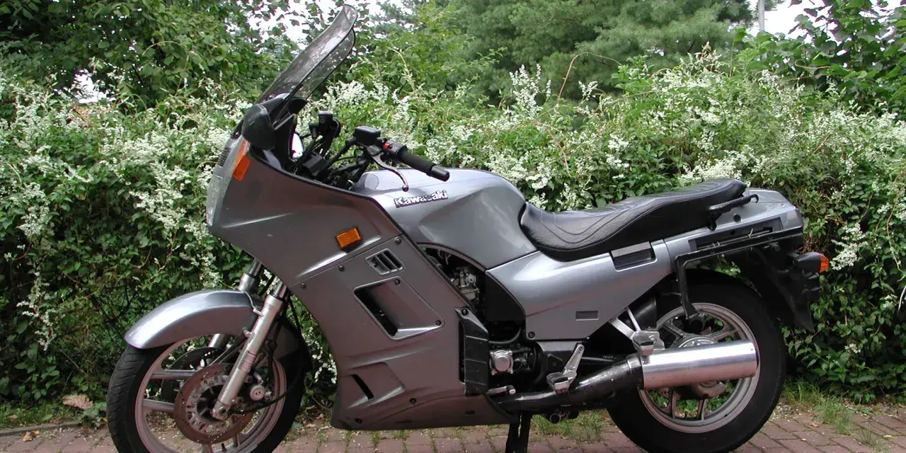 Srebrny motocykl Kawasaki GTR 1000 zaparkowany na czerwonej kostce brukowej, otoczony zielenią.