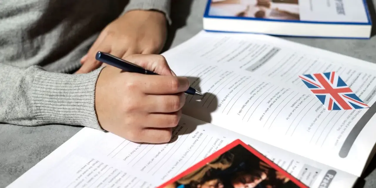 Osoba pisze długopisem w zeszycie z ćwiczeniami z języka angielskiego. Obok leży książka i flaga Wielkiej Brytanii. To może być przygotowanie do napisania klauzuli CV po angielsku.