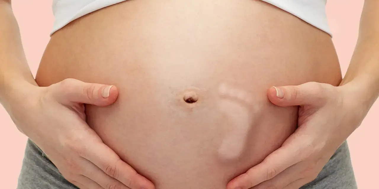 Brzuszek w 30 tygodniu ciąży, z widocznym odciskiem stópki dziecka. Ręce mamy delikatnie obejmują rosnący brzuch.
