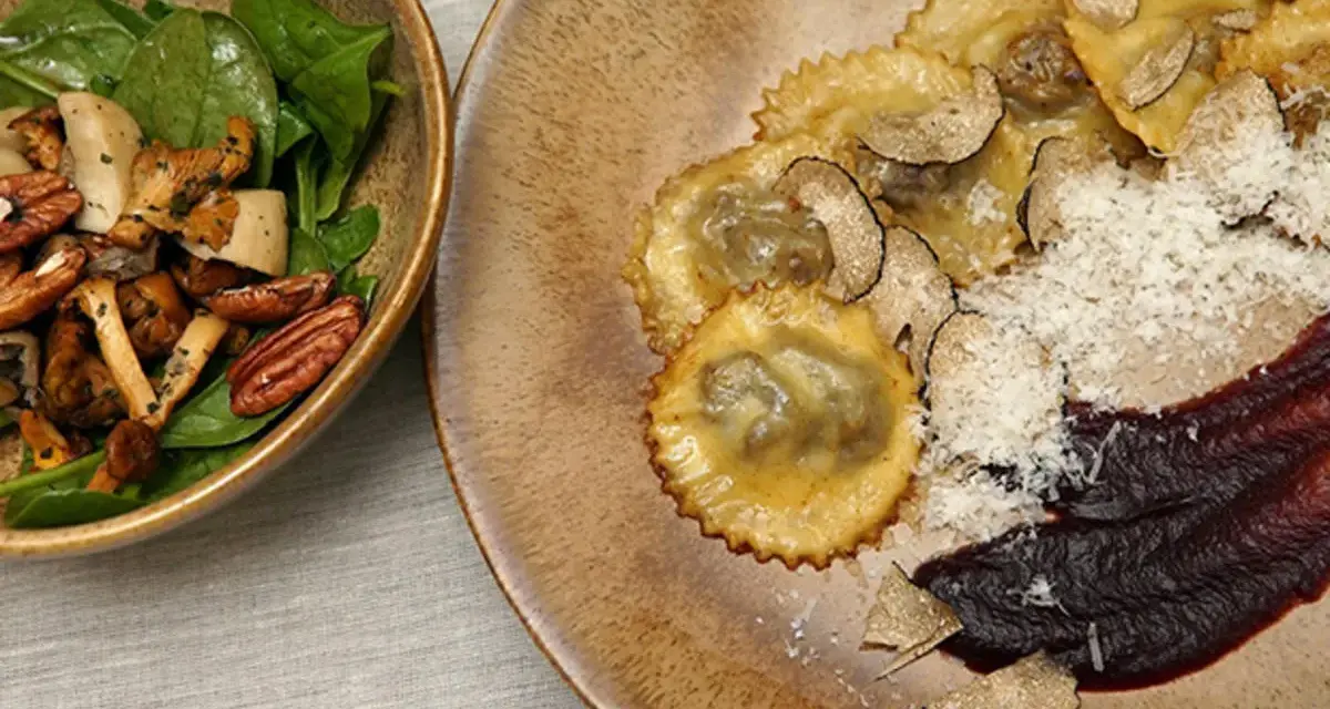 Ravioli pasta mit Trüffeln, Parmesan und einer dunklen Sauce. Daneben eine Schüssel mit Pilzen, Nüssen und Spinat.