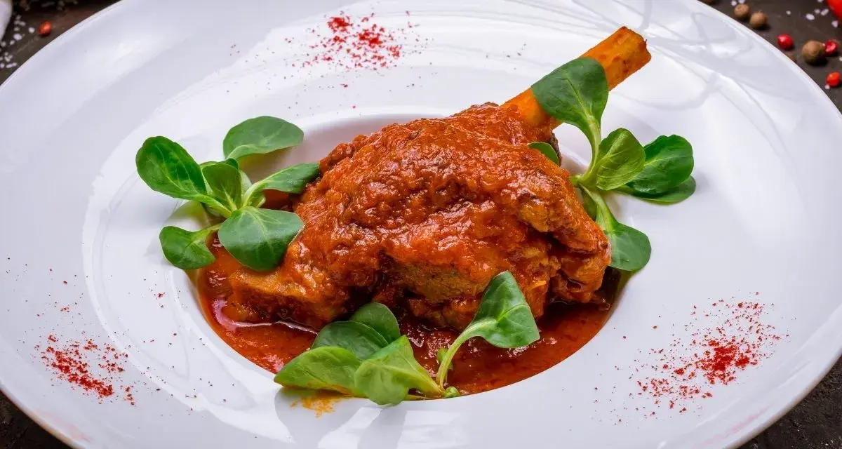 Pierna de cordero en salsa roja con hojas verdes en plato blanco.