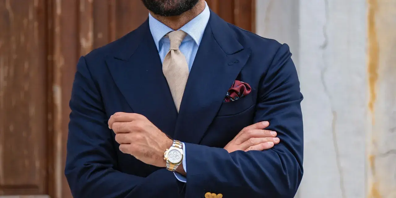 Uomo con abito blu, cravatta beige e fazzoletto bordeaux con un disegno floreale. Il fazzoletto, un dettaglio di stile, aggiunge un tocco di eleganza.