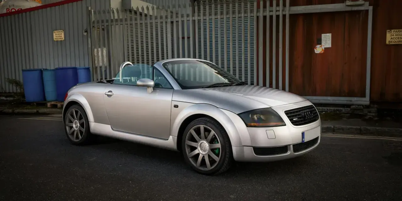 Silberner Audi TT 2002 Roadster mit offenem Verdeck steht auf einer Straße vor einem Industriegebäude.