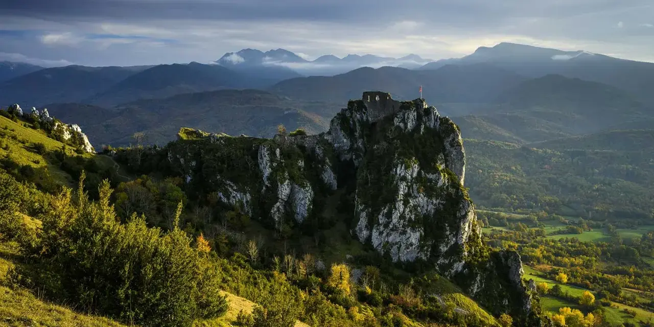 Ruines d'un château cathare sur un pic rocheux, dominant un paysage de montagnes et de forêts. Le chemin des bonshommes serpente à travers la vallée.