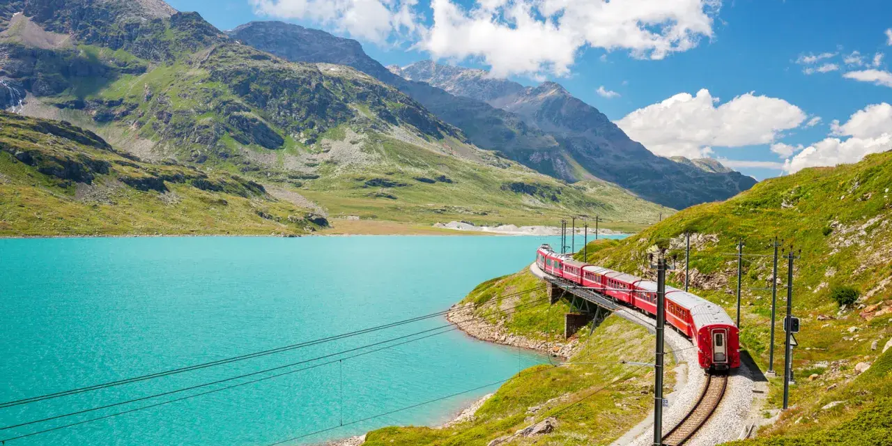 A red train winds along a track beside a vibrant turquoise lake, with lush green mountains and a bright blue sky in the background. This is a classic como to switzerland train journey.