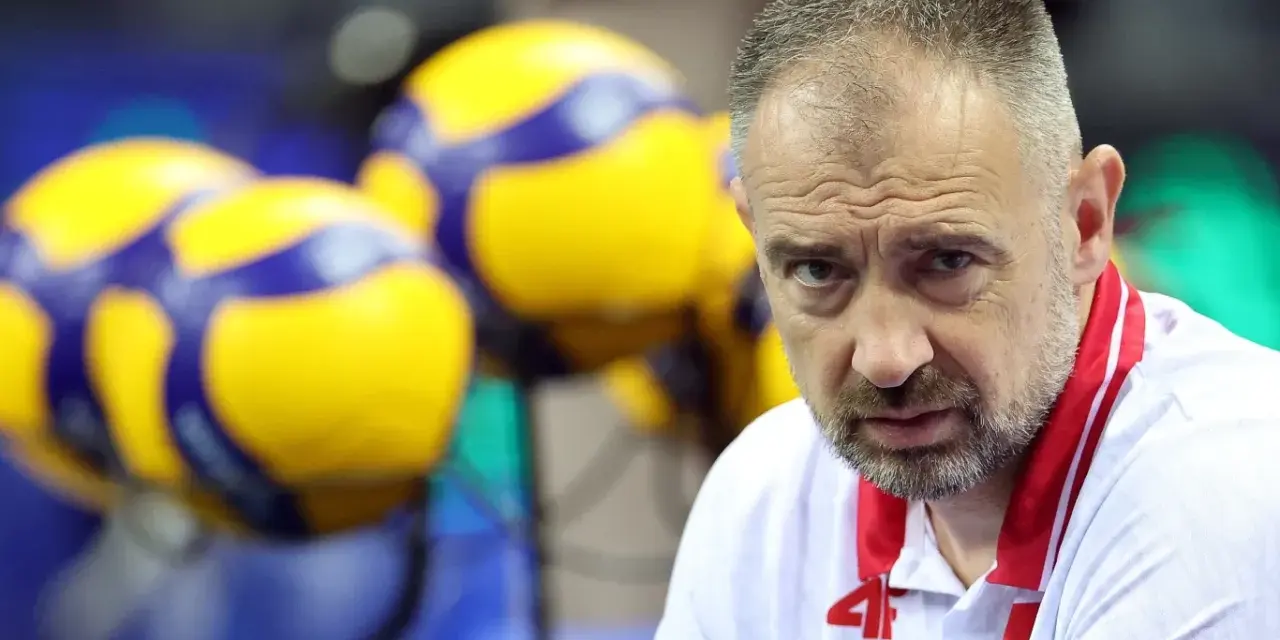 Skupiony Nikola Grbić, trener siatkówki, w białej koszulce z czerwonymi akcentami, na tle żółto-niebieskich piłek.
