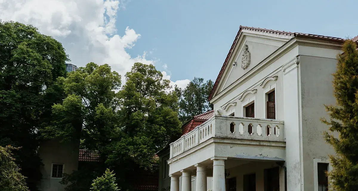 Pałac Grąbkowo, biały budynek z kolumnami i balkonem, otoczony bujną zielenią drzew pod błękitnym niebem z chmurami.