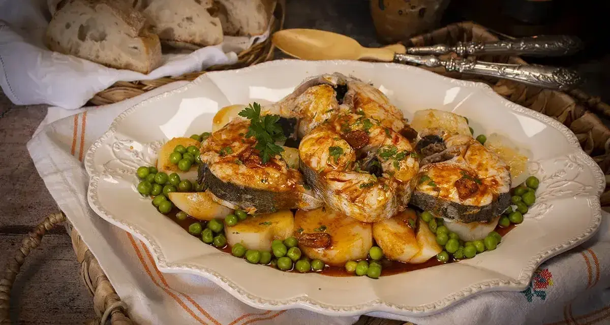 Merluza a la gallega servida en plato blanco con patatas, guisantes y perejil. Pan rústico en cesta al fondo.