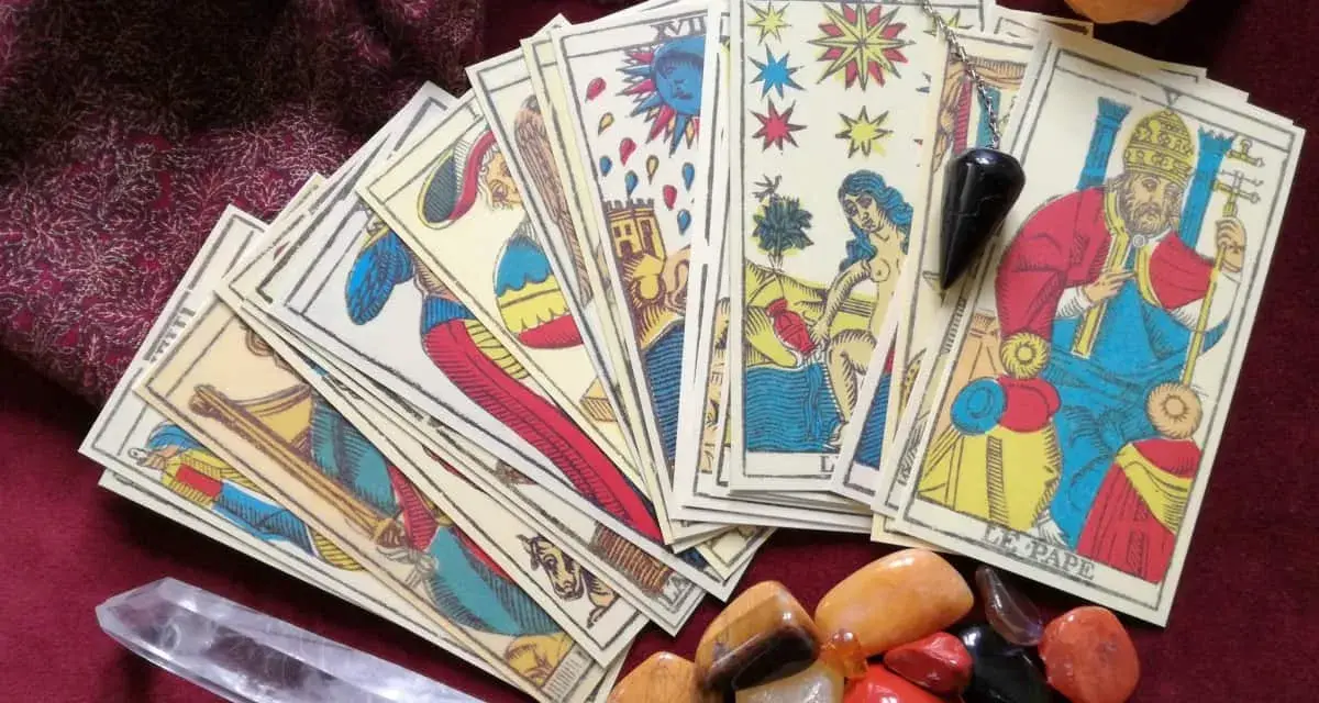 Cartas de tarot psicológico, un péndulo y cristales dispuestos sobre una tela burdeos.