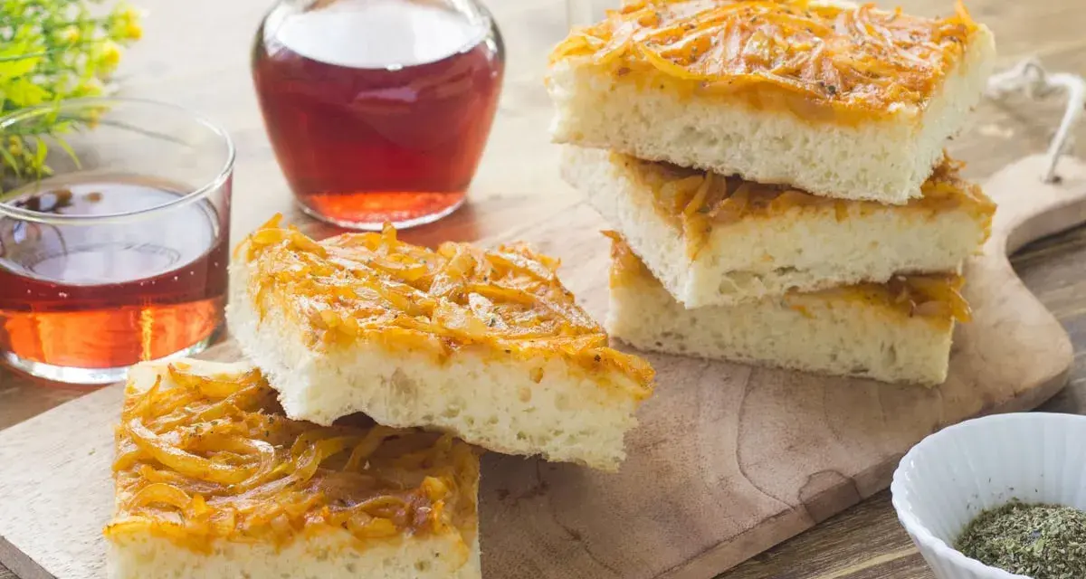 Focaccia con cipolle dorate e soffice, servita su un tagliere di legno con un bicchiere di vino rosato.