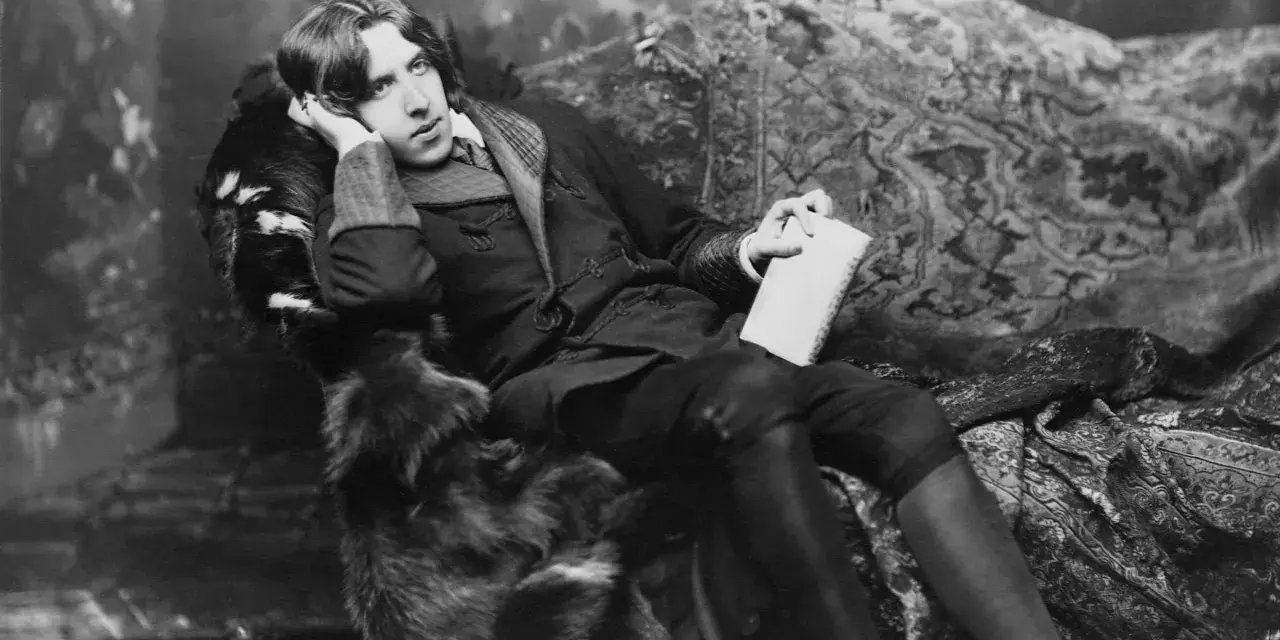 Oscar Wilde, w futrzanym płaszczu, z książką w dłoni, w pozie sugerującej refleksję nad swoimi sztukami.
