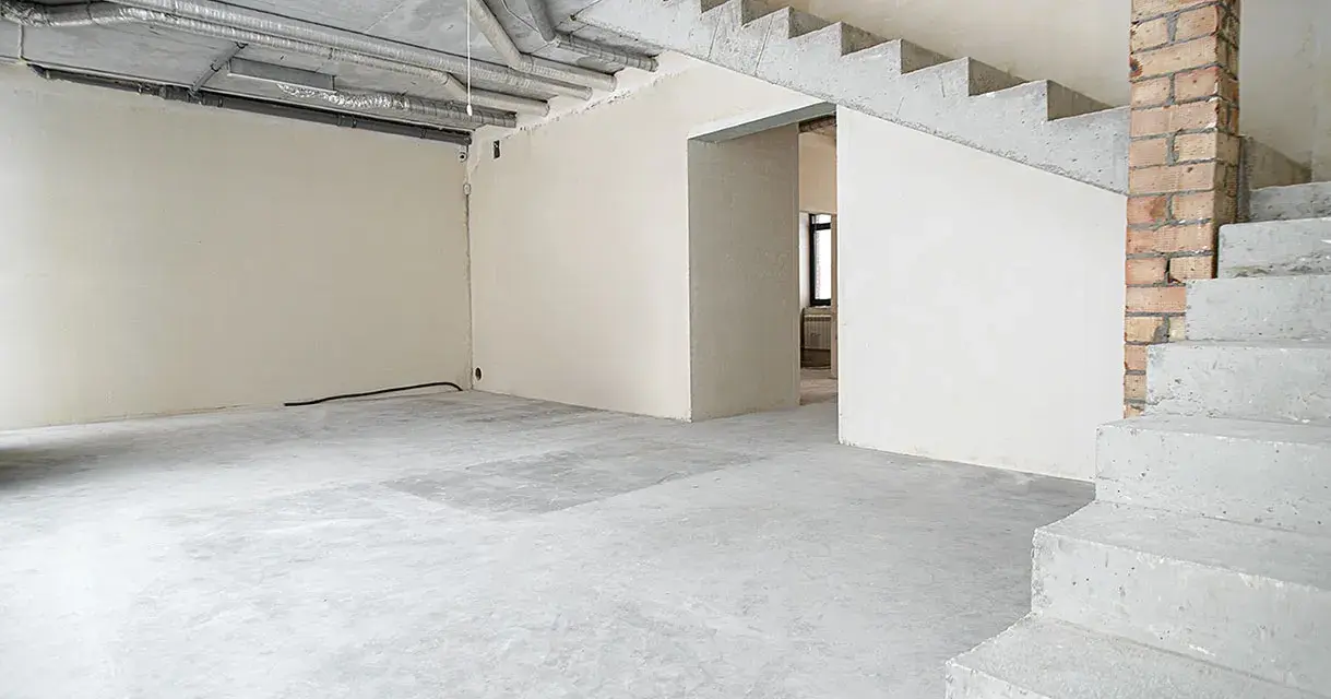 Wnętrze w stanie deweloperskim: betonowe schody, nieotynkowane ściany, widoczne instalacje i przewody.