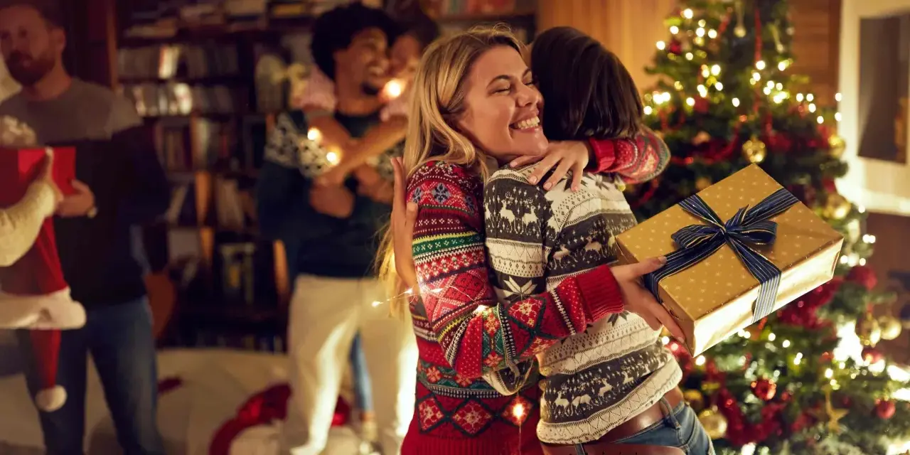Zwei Frauen umarmen sich vor einem Weihnachtsbaum, eine hält ein Geschenk. Eine Familie feiert Weihnachten, eine der vielen schönen weihnachtstraditionen deutschland.