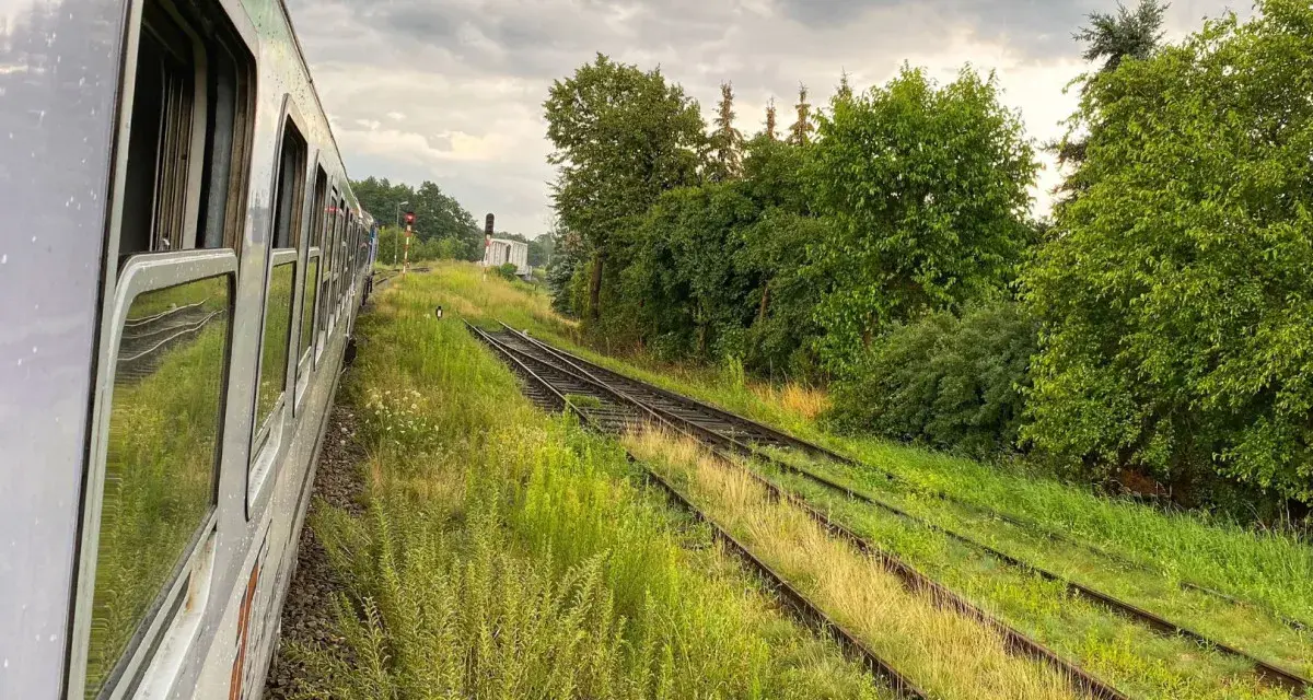 Widok z okna pociągu na tory kolejowe otoczone zielenią i chmury na niebie.