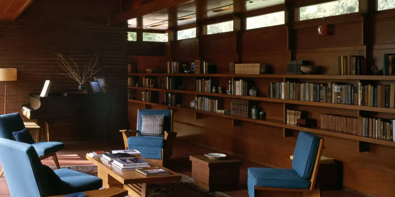 A cozy living room with blue chairs, a piano, and bookshelves. The clerestory windows above let in natural light, illuminating the warm wood interior.