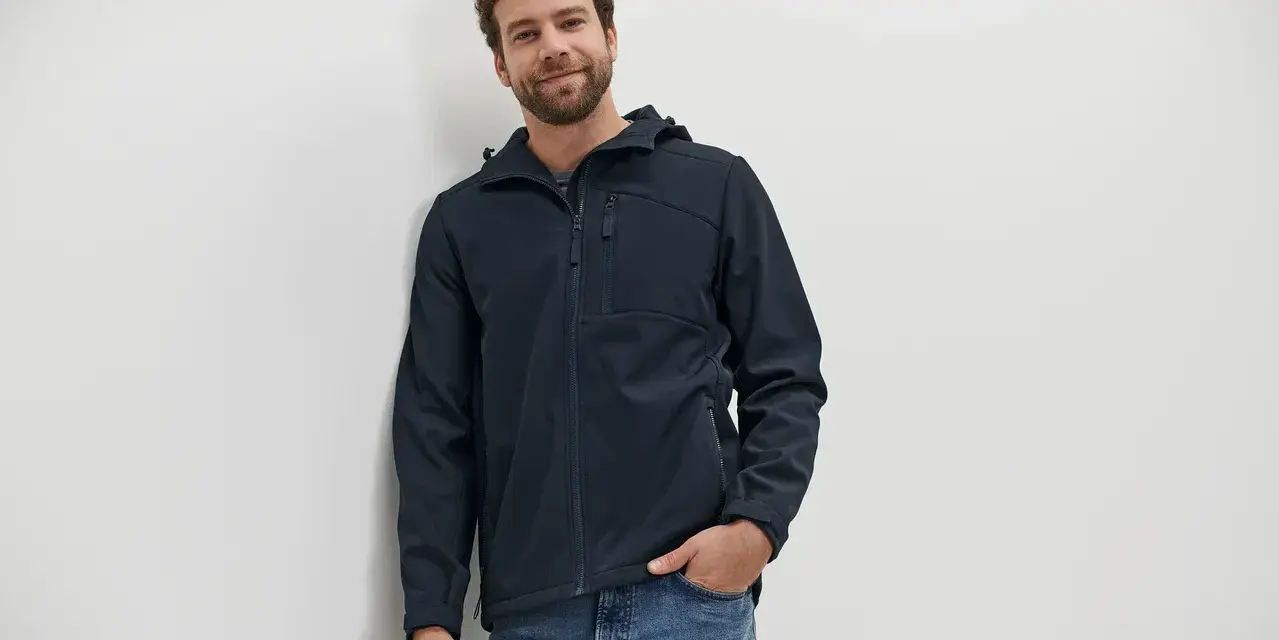 Mann mit lockigem Haar trägt eine dunkelblaue Softshelljacke und Jeans. Perfekt für das nächste Abenteuer oder zum Softshelljacke waschen.