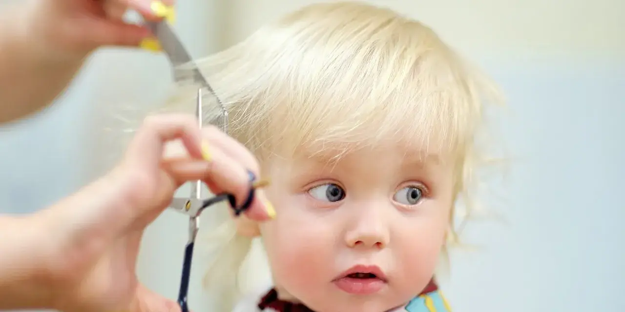 Ein kleines blondes Kind bekommt die Haare geschnitten. Die Schere nähert sich vorsichtig den feinen Strähnen.