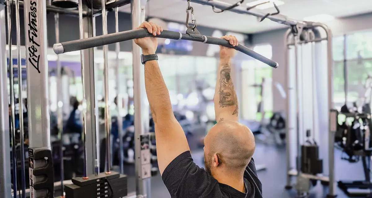 Man med tatueringar utför enarms latsdrag på ett gym.