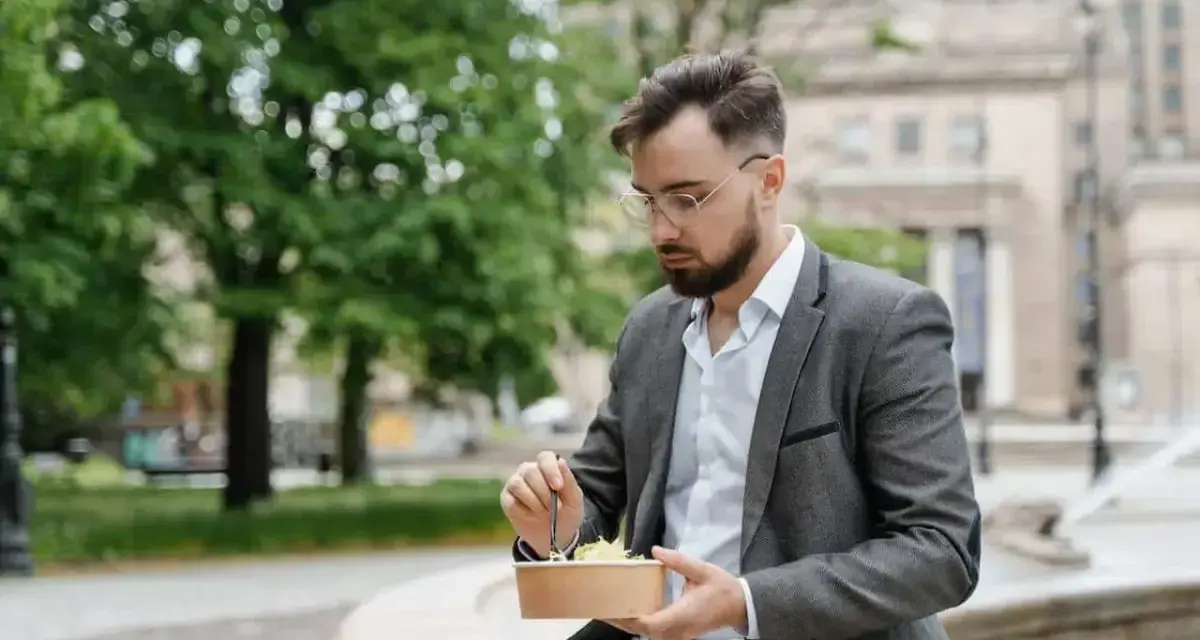 Mężczyzna w okularach i marynarce robi sobie przerwę w pracy, jedząc sałatkę na ławce w parku.