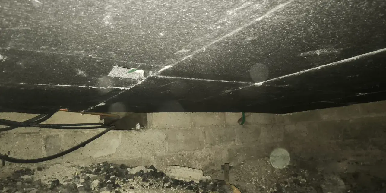 Vue de dessous d'un plancher, montrant le matériau d'isolation du plancher bas, des tuyaux et des blocs de béton.