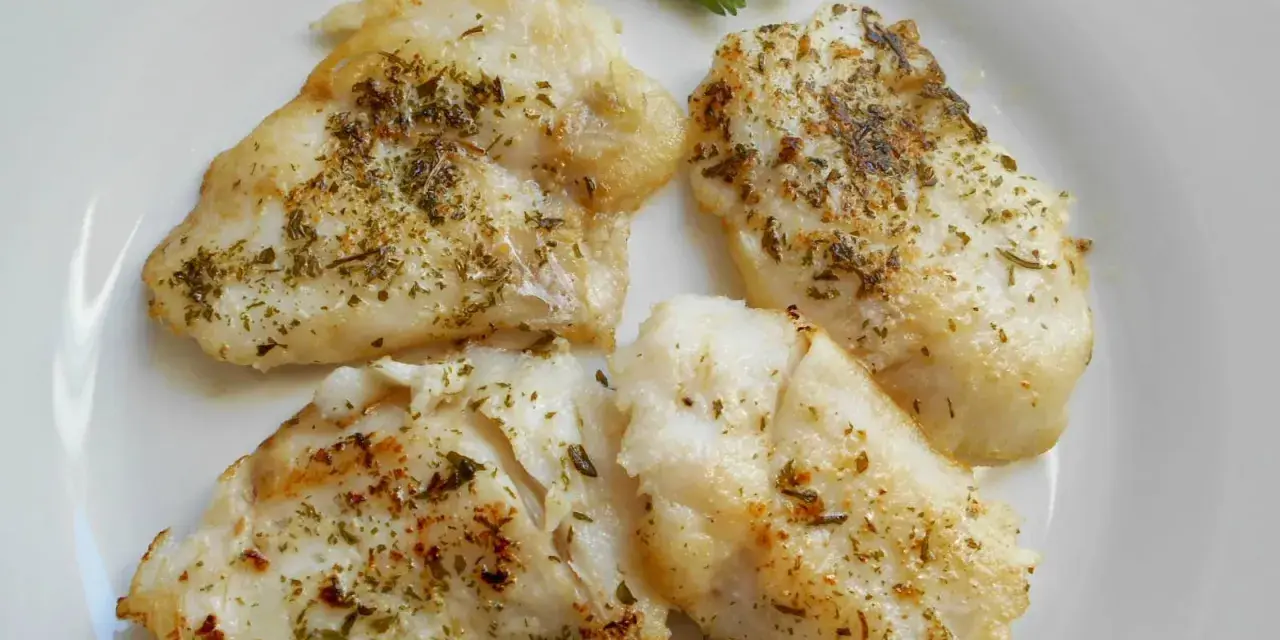 Cuatro trozos de pescado blanco rosada a la plancha, sazonados con hierbas, servidos en un plato blanco con una ramita de perejil.
