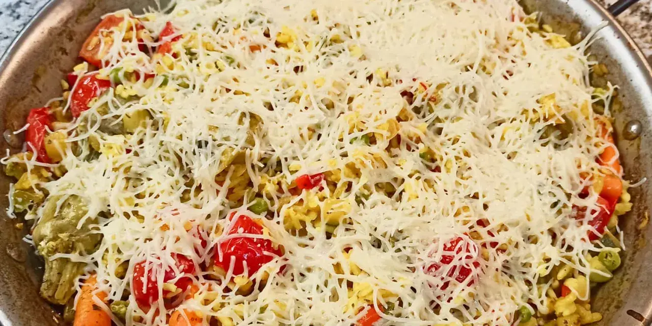 Arroz a la milanesa listo para hornear, cubierto con queso rallado y vegetales frescos como tomates, zanahorias y guisantes.