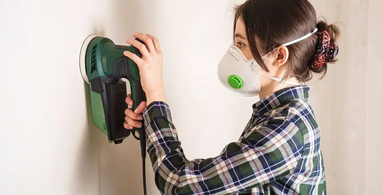 Persona con mascarilla y camisa a cuadros usa una lijadora eléctrica para lijar una pared.