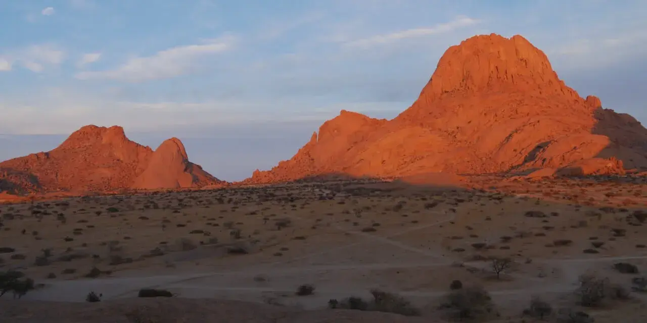 Zachód słońca maluje na pomarańczowo skalne szczyty Spitzkoppe w Namibii. Pustynny krajobraz z suchymi krzewami i ścieżkami.