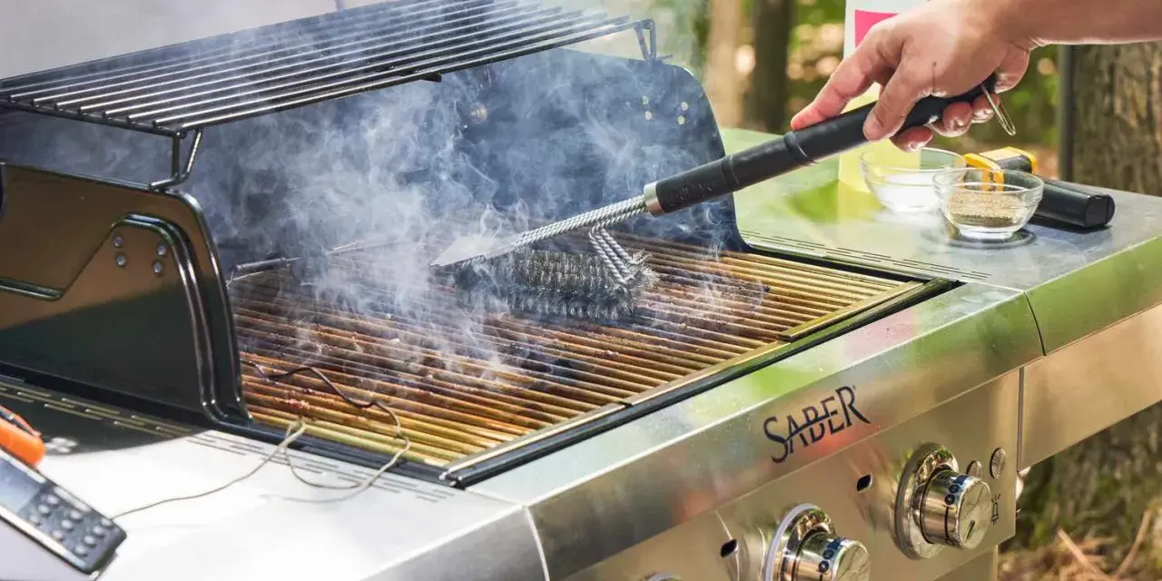 A hand uses a grill brush to clean the grates of a smoking Saber grill, showing the best way to clean a grill for your next cookout.