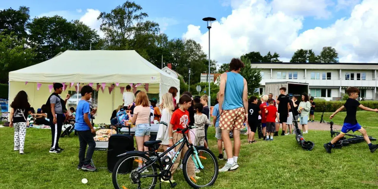 Barn och vuxna samlas vid ett tält på en gräsmatta, kanske för att dela ett hemligt språk. En cykel och elsparkcyklar syns.
