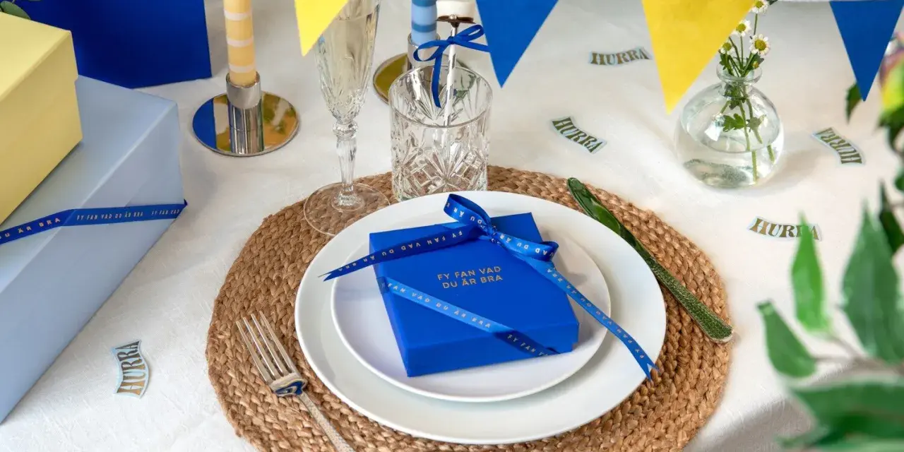 Blå och gula girlanger, ljus och presenter på ett dukat bord. En blå present med band och texten "Fy fan vad du är bra" är en fin pom pom dekoration.