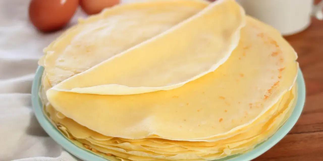 Piatto di crepes senza burro, leggere e dorate, con due uova sullo sfondo.