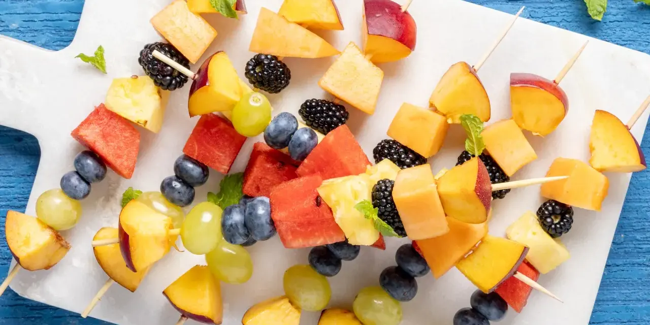 Spiedini di frutta colorati con pesche, melone, anguria, ananas, mirtilli, more e uva, guarniti con menta fresca.