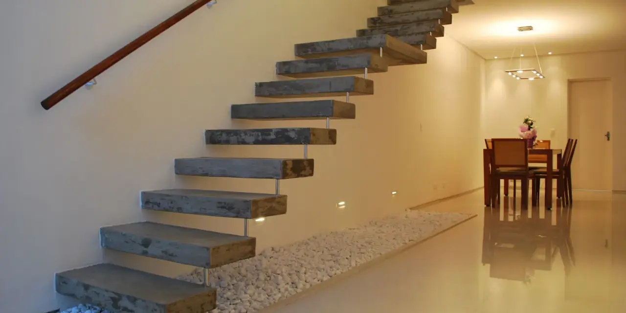 Escaleras rústicas modernas de hormigón flotante con pasamanos de madera. Un camino de guijarros blancos bordea la base.