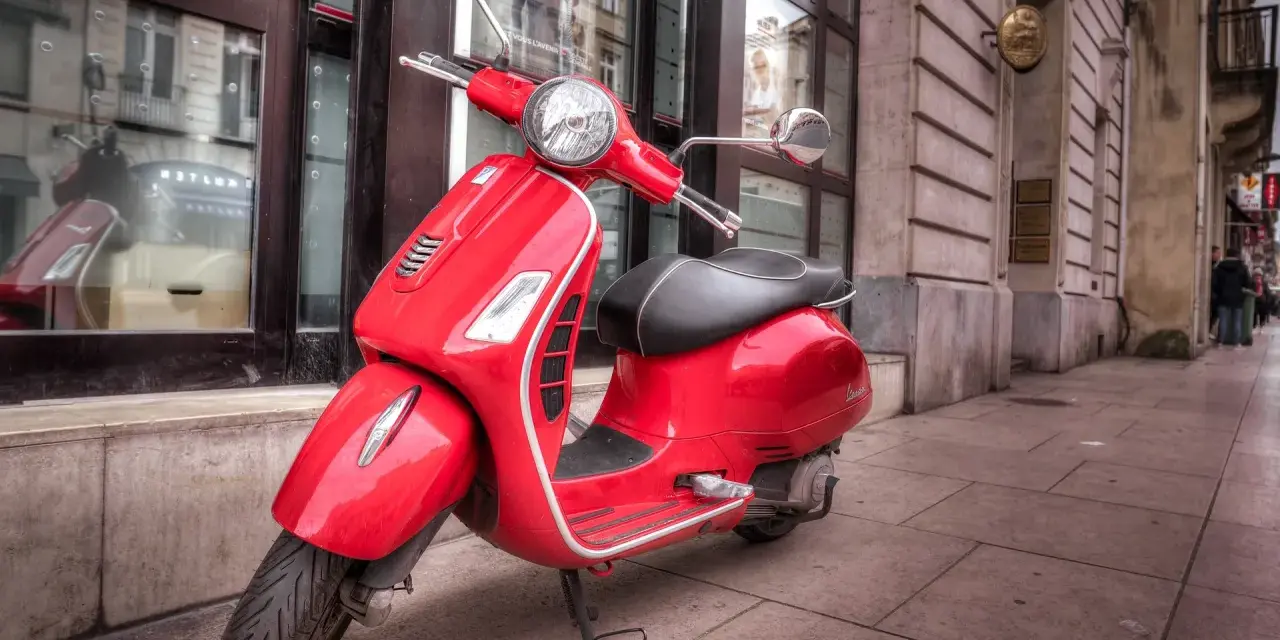 Czerwony skuter Vespa, idealny do zdobycia prawa jazdy AM, stoi na chodniku przed budynkiem.