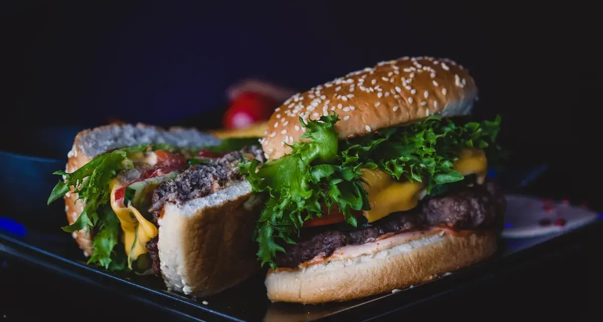 Dwa soczyste burgery z serem, sałatą i warzywami na ciemnym tle.