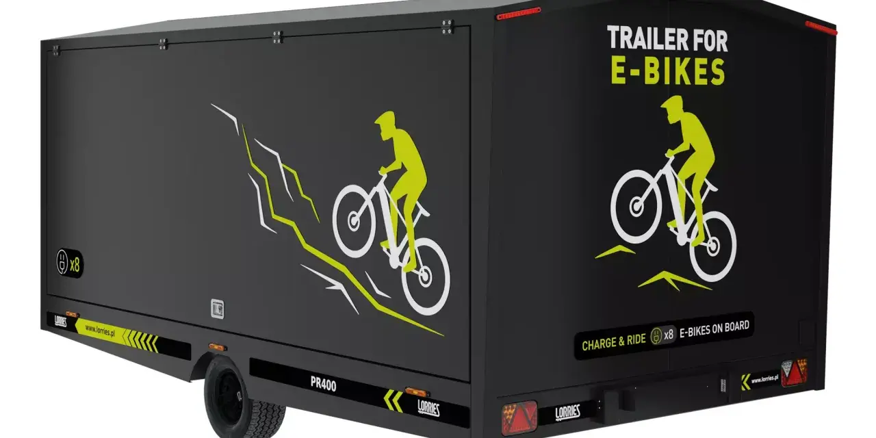Czarna rowerowa przyczepa kempingowa z grafiką rowerzysty na górskim szlaku. Napis "TRAILER FOR E-BIKES" i "CHARGE & RIDE x8 E-BIKES ON BOARD".