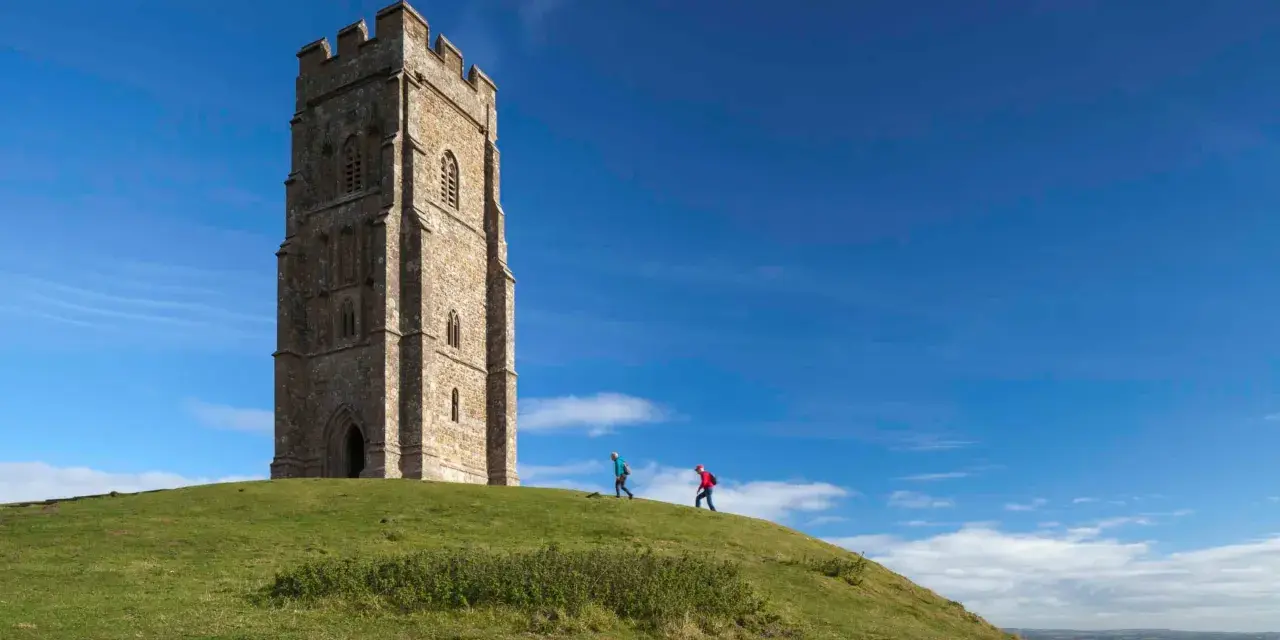 Two hikers ascend the grassy slope towards the iconic Glastonbury Tor tower under a bright blue sky.