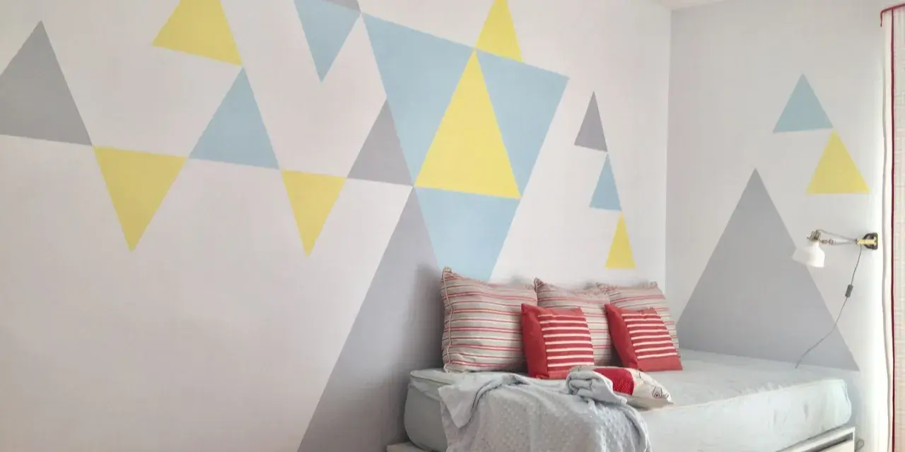 Pared pintada con triángulos de colores pasteles, creando un diseño geométrico moderno. La habitación tiene una cama con cojines y una manta.
