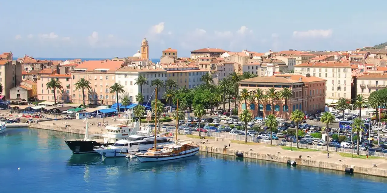 Malowniczy port na wyspie z Ajaccio, z luksusowymi jachtami i palmami, w tle kolorowe kamienice i wieża kościelna.