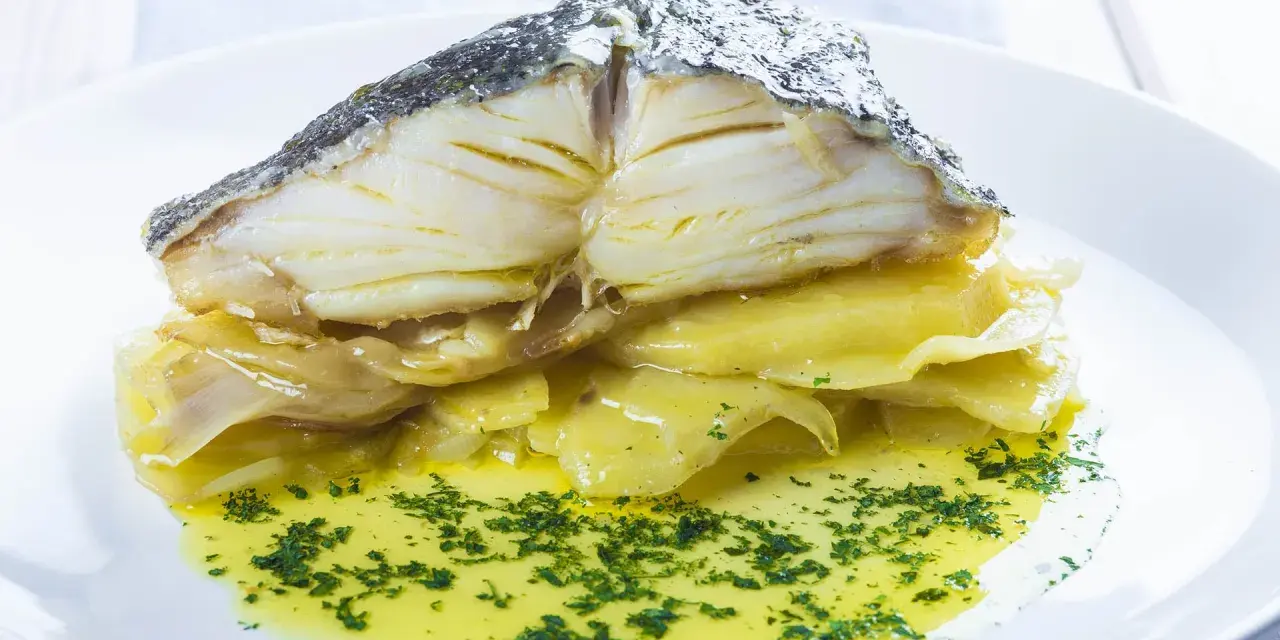Merluza al horno con patatas, jugosa y dorada, servida sobre patatas tiernas y bañada en aceite de oliva con perejil picado.