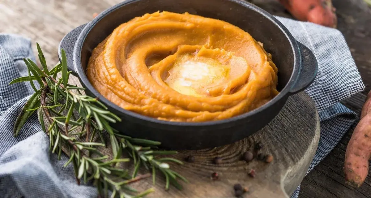 Un cremoso puré de zanahoria en un cuenco negro, coronado con mantequilla derretida. Romero fresco y granos de pimienta añaden aroma.