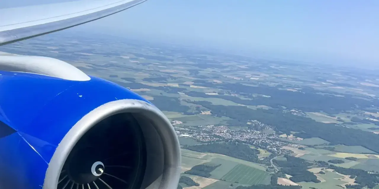 View from an airplane window, showing a blue engine and wing, with a patchwork of green fields and a small town below. Perfect for day trip flights.