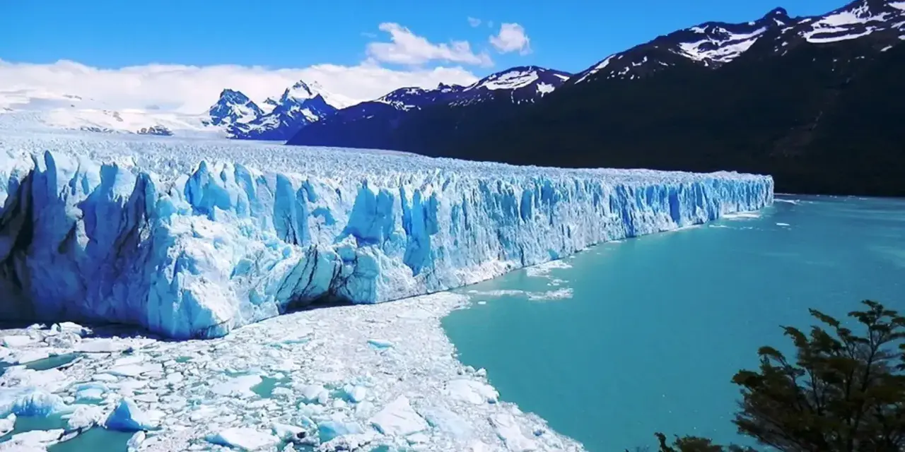 Impresionante glaciar Perito Moreno, con sus paredes de hielo azul intenso y el agua turquesa del lago. Montañas nevadas al fondo.