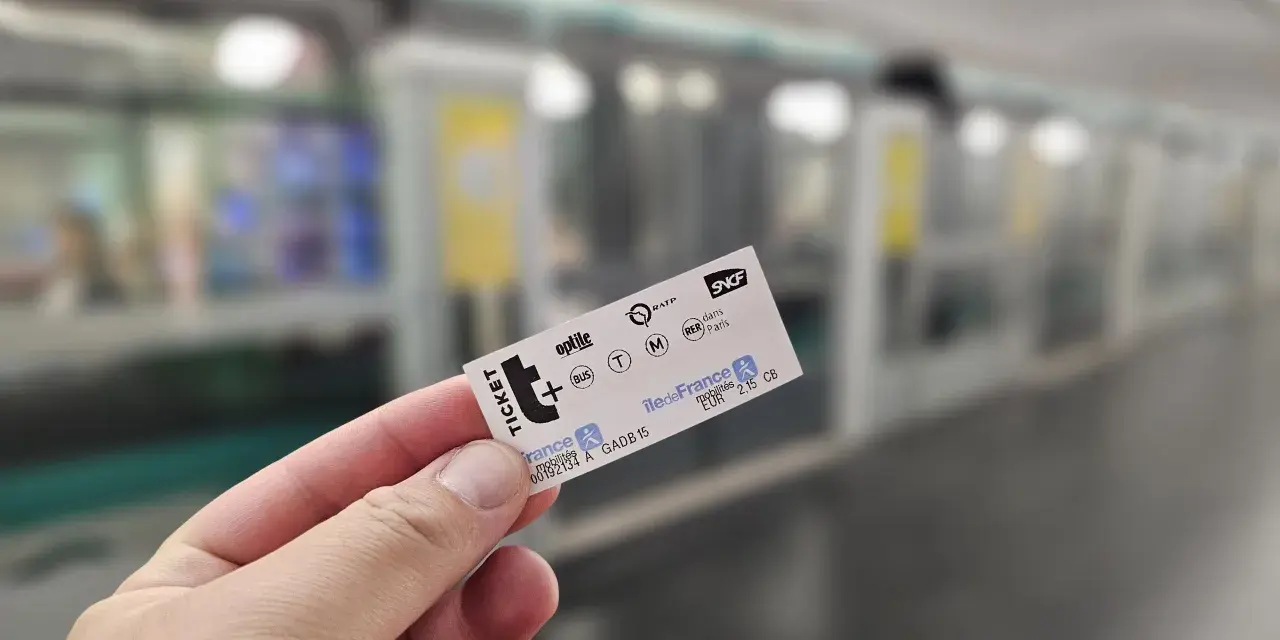 Mano sostiene un ticket t+ para el transporte público en París, con un tren borroso al fondo.