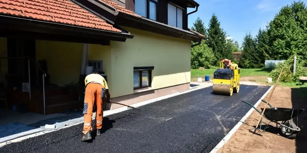 Pracownik równa łopatą świeży asfalt na podjazd, a walec go ubija. Dom w tle.