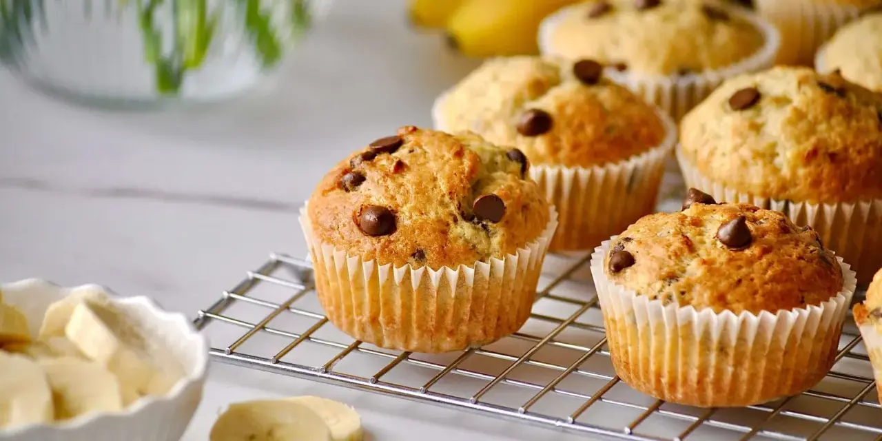 Frisch gebackene bananen muffins mit Schokostückchen auf einem Gitter, daneben eine Schüssel mit Bananenscheiben.