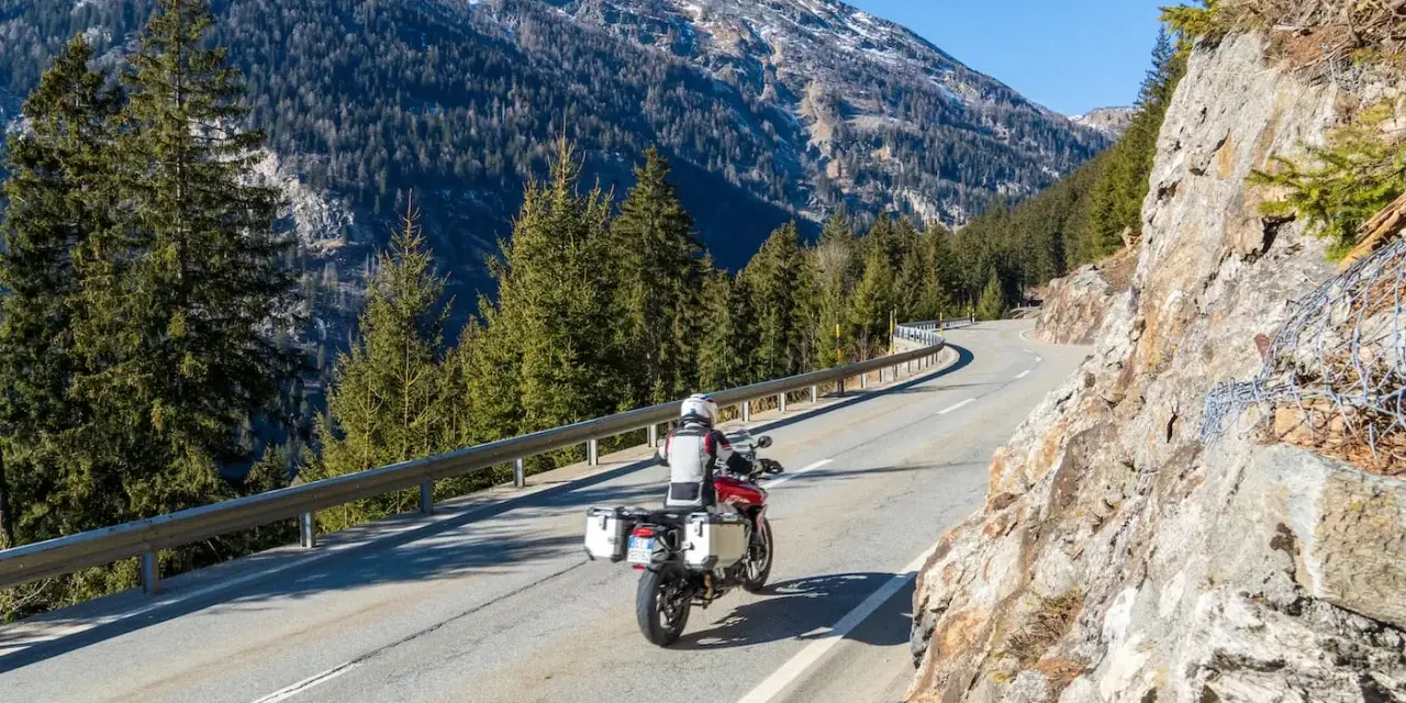Motociclista su strada alpina in Svizzera, con montagne innevate e foreste. Ottimi consigli per un viaggio in moto in Svizzera.